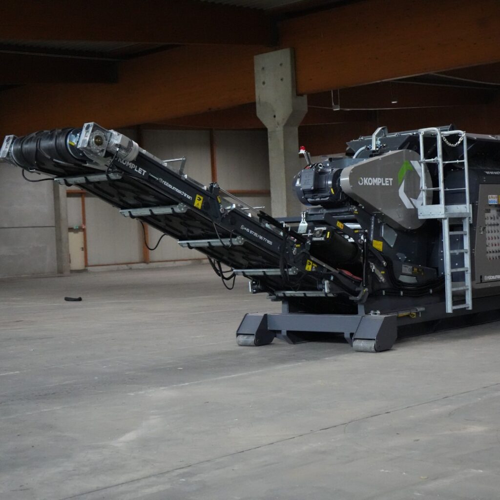 Ein grauer KOMPLET-Mobilbrecher mit langem Förderband steht in einer Halle, während die Anlage und ihre Metallkonstruktion deutlich zu sehen sind.