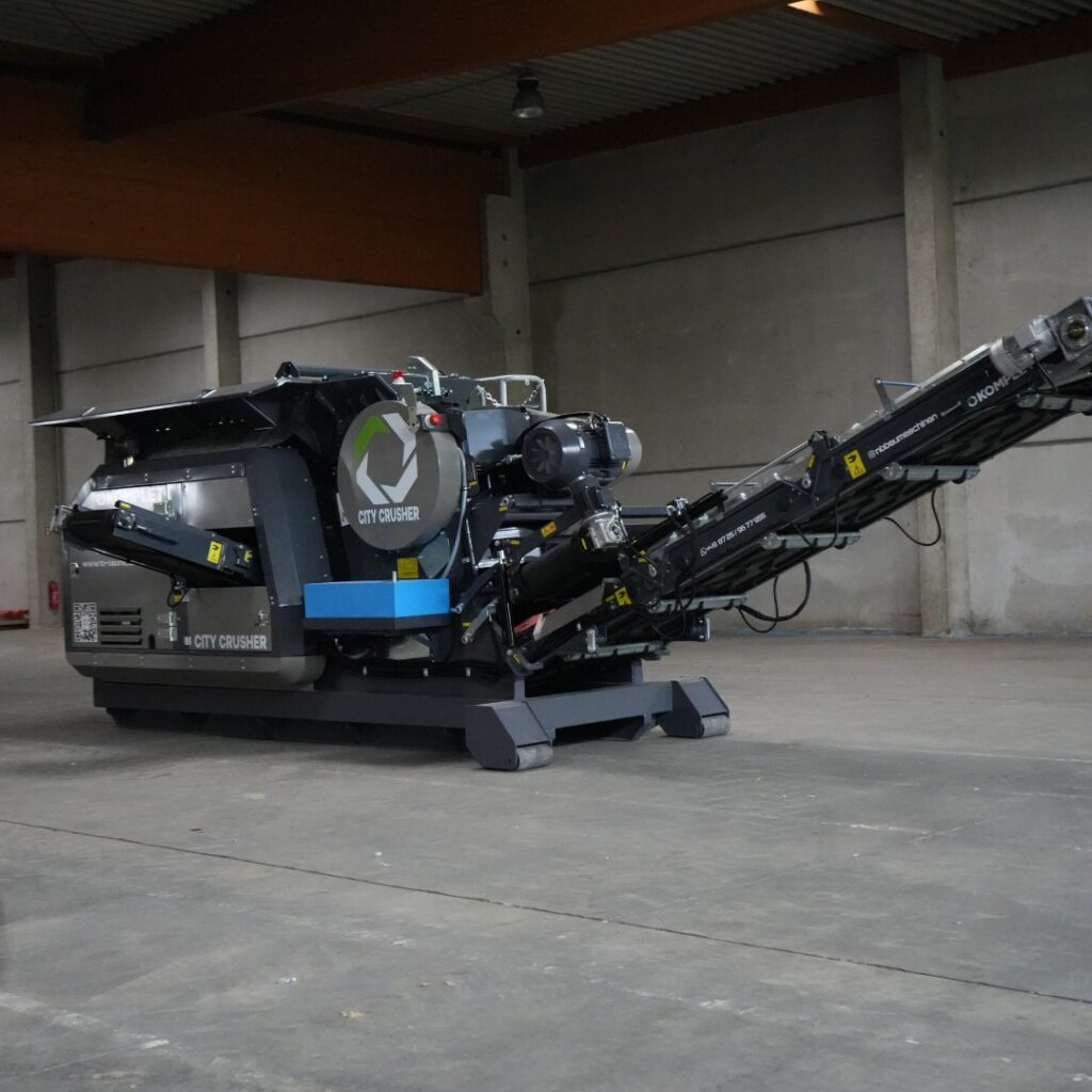 In einer großen Halle steht ein schwarzer City Crusher mit langem Förderband, markantem Rundlogo und robuster Technik auf dem Betonboden.