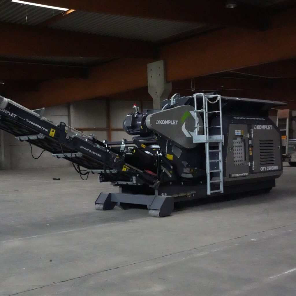 In einer Halle steht ein dunkelgrauer Komplet City Crusher mit Förderband, Leiter und Bedieneinheit auf einem Betonboden unter Stahlträgern.