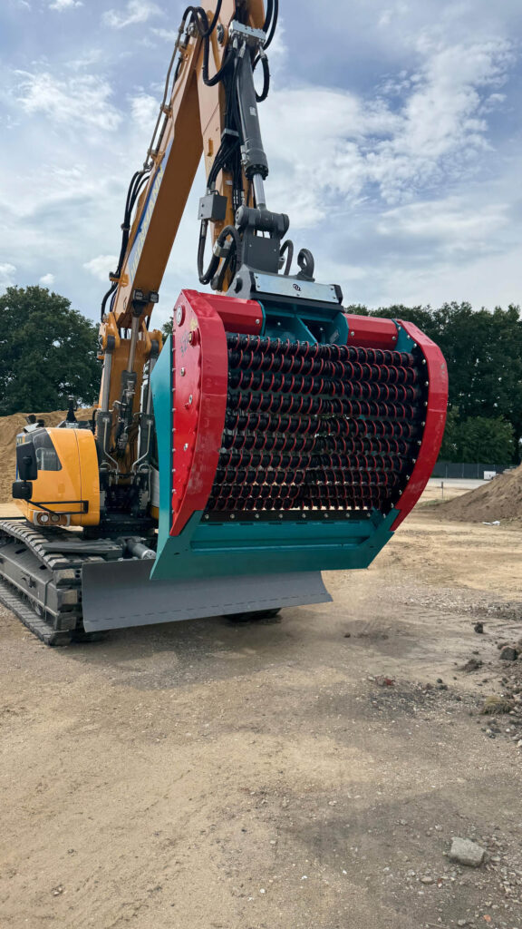 Ein gelber Kettenbagger trägt einen rot-türkisen Sieblöffel mit vielen schwarzen Drehspiralen auf einer Baustelle unter wolkigem Himmel.
