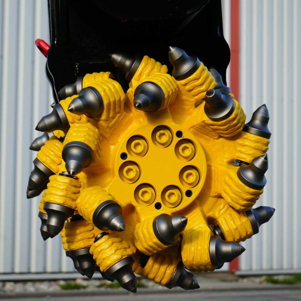 Ein gelb-schwarzer Fräskopf mit vielen konischen Spitzen hängt vor einer hellen Metallwand und zeigt die massiven Schneidwerkzeuge im Detail.