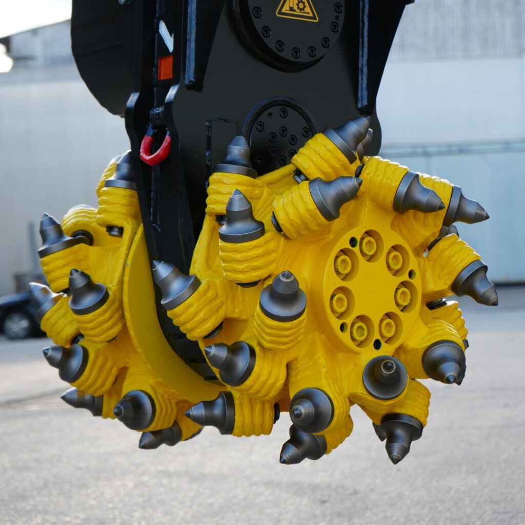 Ein gelb-schwarzer Fräskopf mit vielen Hartmetallspitzen hängt vor einem hellen Industriegelände, mit sichtbaren Hydraulikdetails und Metallgehäuse.