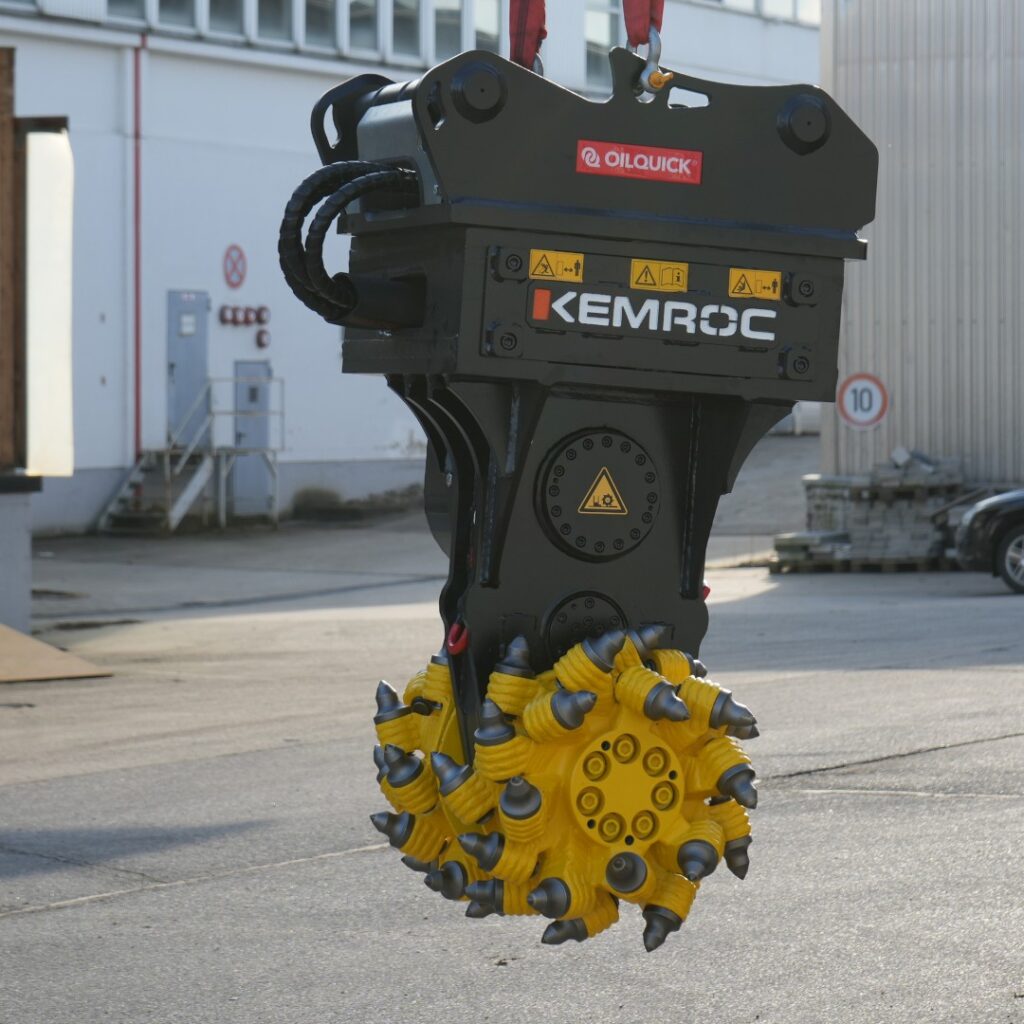 Ein schwarzer KEMROC-Fräskopf mit gelber Walze und OILQUICK-Aufhängung hängt vor einer Industriehalle im Tageslicht auf grauem Asphalt.