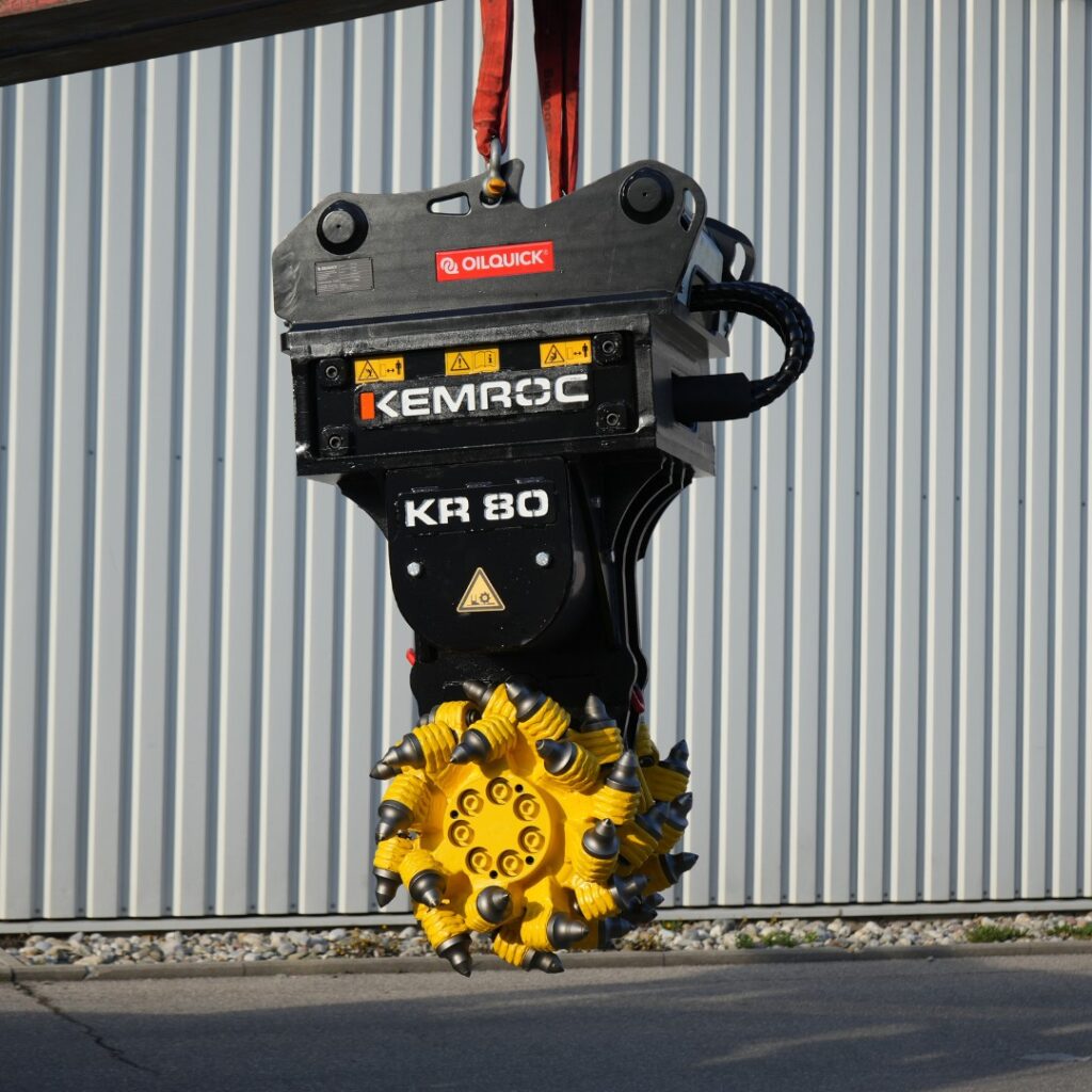 Ein schwarzer KEMROC KR 80 mit OILQUICK-Aufnahme und gelbem Fräskopf hängt vor einer hellen Metallfassade, während rote Hebegurte sichtbar sind.