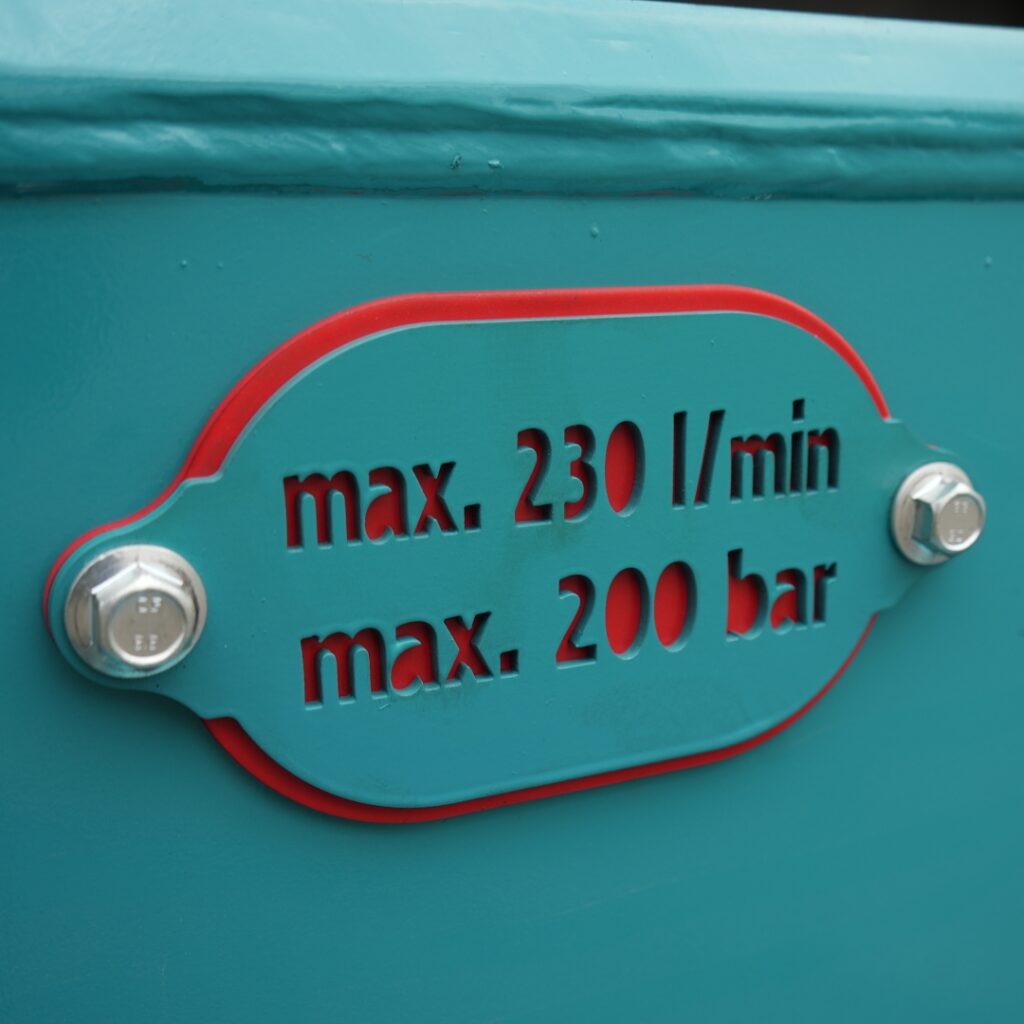 Eine türkisfarbene Metallplakette mit roter Umrandung zeigt die Angaben „max. 230 l/min“ und „max. 200 bar“, befestigt mit zwei Schrauben.