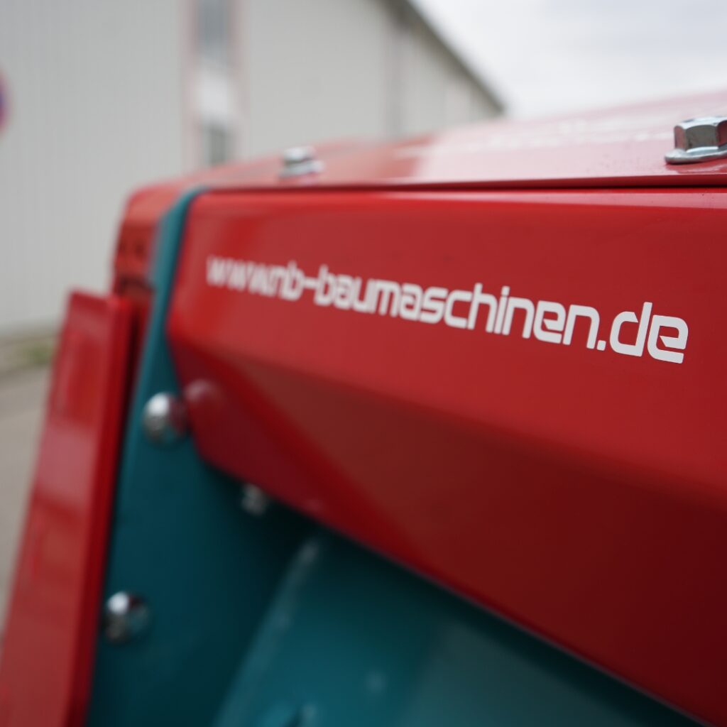 Nahaufnahme eines roten Maschinengehäuses mit weißem www.nb-baumaschinen.de-Schriftzug und türkisfarbener Innenfläche vor unscharfem Hintergrund.