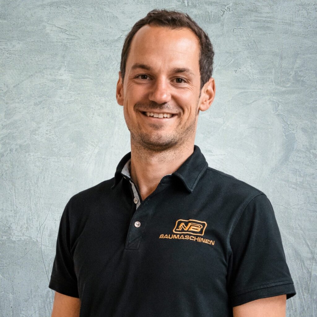 Ein lächelnder Mann im schwarzen NB-Baumaschinen-Poloshirt steht vor einem hellgrau strukturierten Hintergrund und blickt direkt in die Kamera.