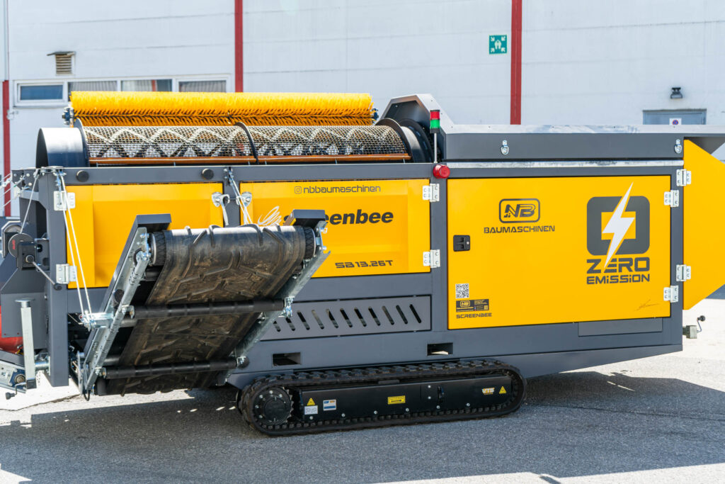 Eine gelb-graue mobile Trommelsiebanlage auf Kettenfahrwerk steht vor einer Industriehalle; oben sind Bürsten, seitlich steht Zero Emission.