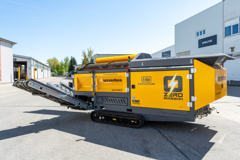 Gelbe SCREENBEE SB13.26e-Track mit Raupenfahrwerk und Förderschnecke steht vor Industriehallen, daneben sind NB Baumaschinen und Zero Emission zu sehen.