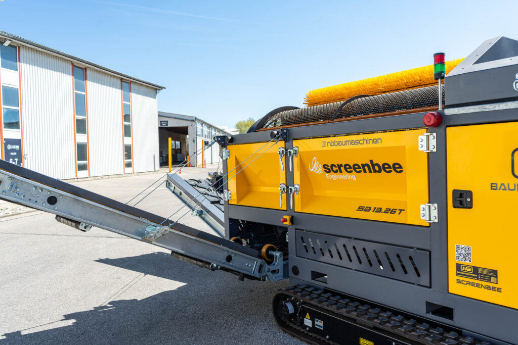 Gelbe Trommelsiebanlage Screenbee SB13.26T auf Raupenfahrwerk neben einem Industriegebäude, mit Förderband, Bürste und Warnleuchte im Freien.