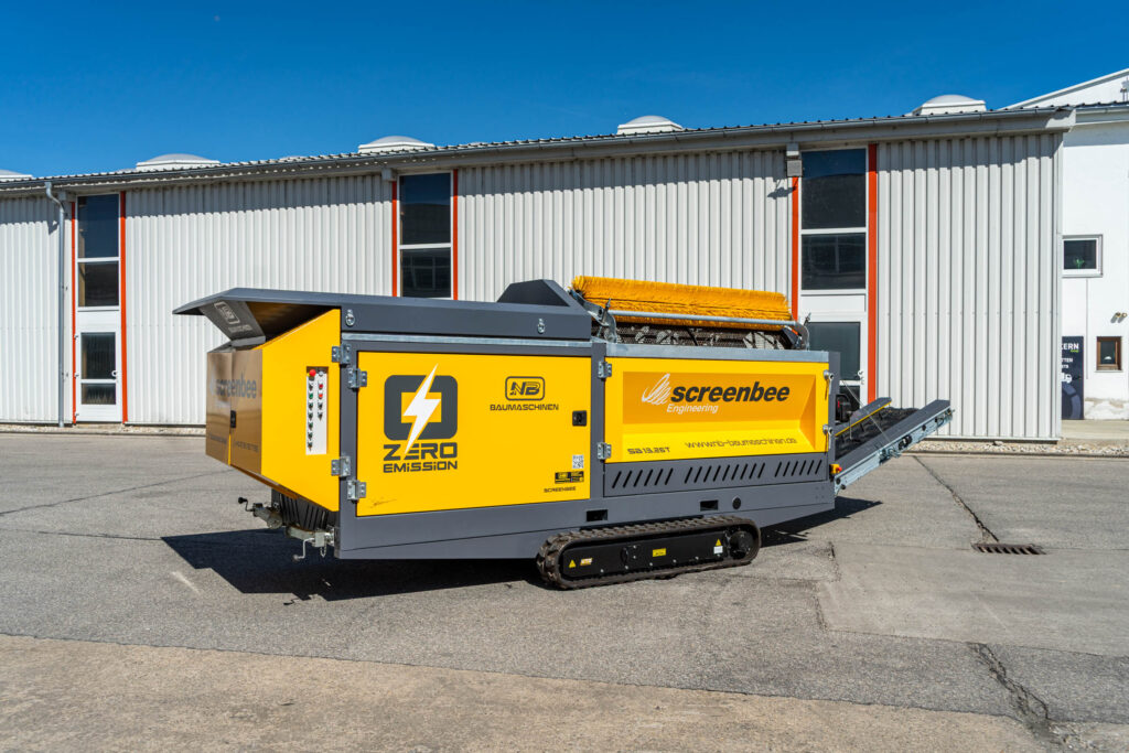 Die gelbe Screenbee SB13.26e-Track von NB Baumaschinen steht auf Raupenfahrwerk vor einer Hallenfassade mit blauem Himmel und klarem Licht.