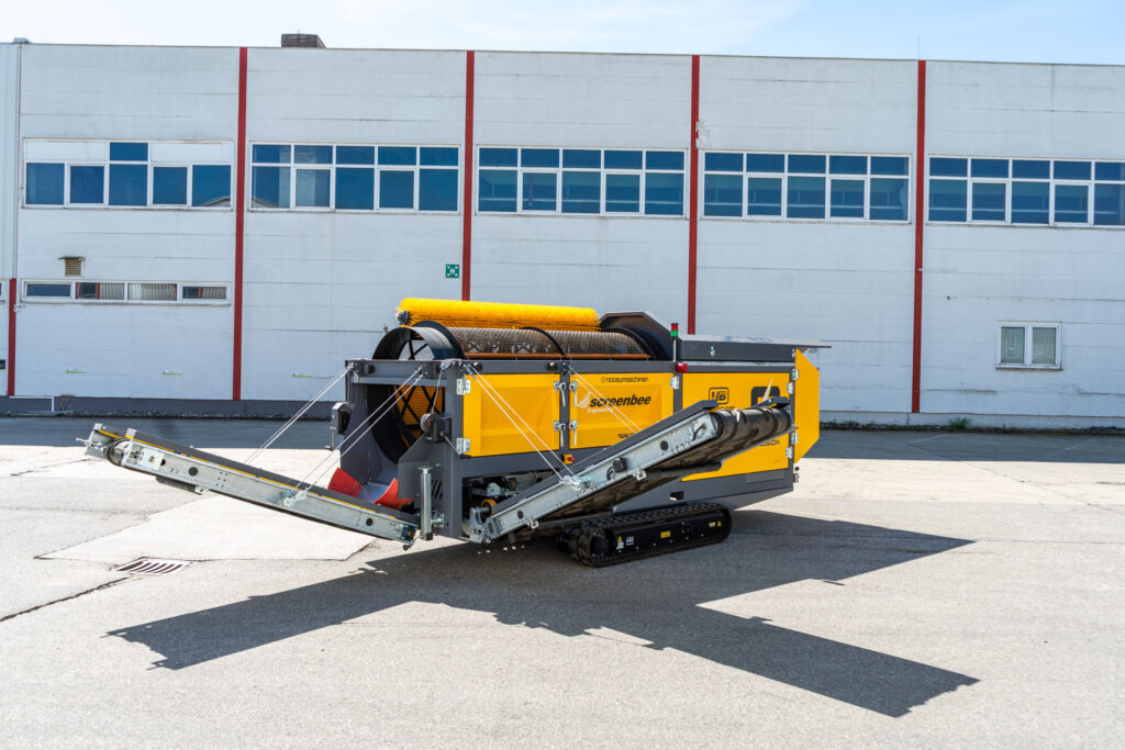Gelbe Screenbee SB13.26e-Track mit Kettenfahrwerk, Trommel und Förderbändern steht vor einer weißen Industriefassade auf Asphalt.