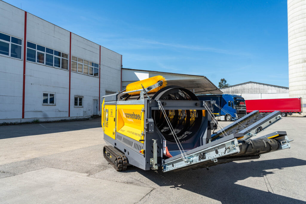 Eine gelb-schwarze Screenbee SB13.26e-Track mit Raupenfahrwerk steht vor Industriehallen, daneben sind Förderband, Trommel und Bürste sichtbar.