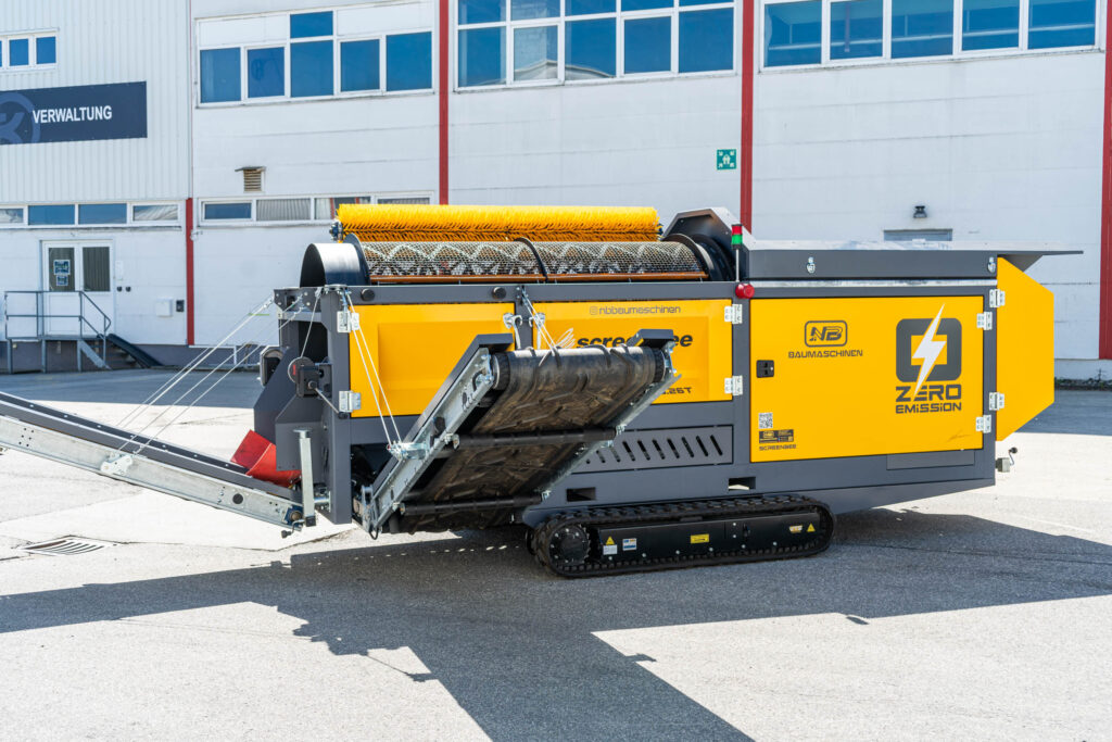 Gelbe Trommelsiebanlage Screenbee SB13.26e-Track von NB Baumaschinen mit Kettenfahrwerk, Bürstenwalze und Förderband vor einem Verwaltungsgebäude.